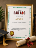 A gold-framed novelty certificate reading 'Official BAD ASS OF THE YEAR AWARD' displayed on a wooden surface,  and a rolled diploma tied with a red ribbon in front.